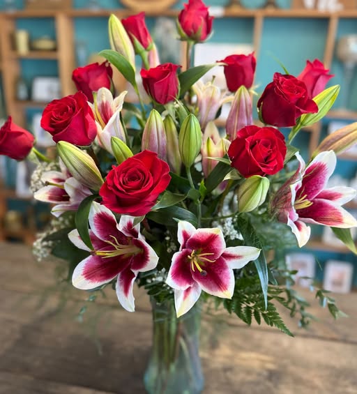 Bouquet of red roses and pink‑white lilies in a glass vase on a wooden table, with a soft blurred background.