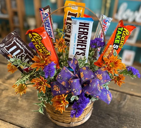 Halloween-themed gift basket with assorted candy bars (Hershey's, Snickers, Reese's, Twix) and orange/purple flowers in a wicker container.