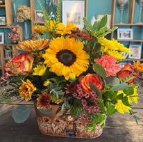 Basket containing a bright bouquet of sunflowers, roses, and assorted flowers in a woven container on a table.