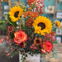 Vibrant bouquet featuring yellow sunflowers, orange roses, and pink blossoms in a white vase.